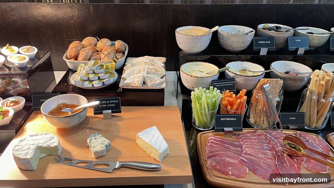 A buffet spread featuring a variety of appetizers and charcuterie. In the foreground, there are cheeses, jam, and sliced cured meat. Behind these, there are bread rolls, pita bread, dips, vegetable sticks (celery and carrots), breadsticks, and olives.