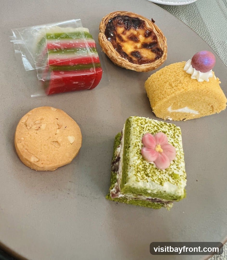 A plate with a variety of small desserts, including a layered jelly, a Portuguese egg tart, a slice of Swiss roll cake, a cookie, and a matcha layered cake decorated with a pink flower.