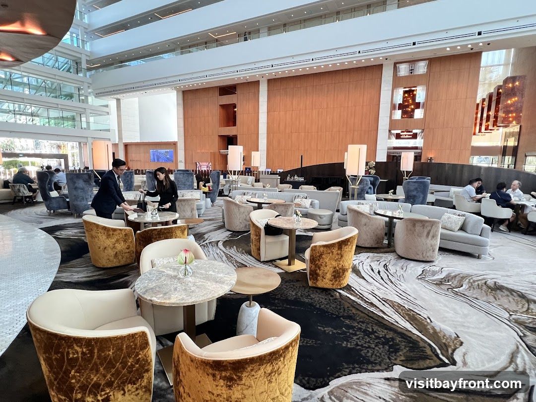 A spacious and elegantly designed hotel lobby lounge with plush seating arrangements, including velvet armchairs and sofas, arranged around marble-topped tables. The space features a high ceiling with modern lighting fixtures and large windows, creating a bright and airy atmosphere. Several guests are seated, some interacting with staff, while others relax in the comfortable seating. The floor is covered with a large, abstract patterned carpet.