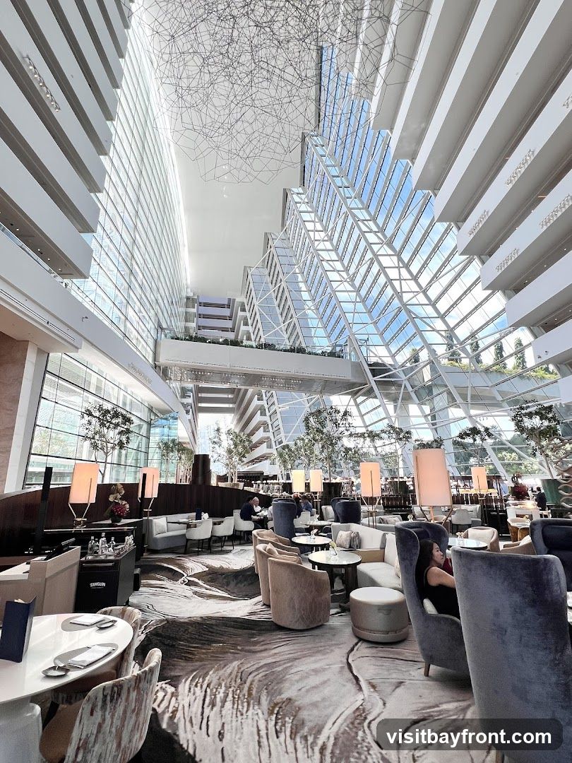 An interior view of a spacious, modern lounge area within a large atrium. The architecture features soaring glass walls and a geometric ceiling structure, allowing natural light to flood the space. Comfortable seating arrangements, including plush armchairs and sofas, are scattered throughout the room, some around small tables. Potted trees and elegant lamps add to the ambiance. Several patrons are seated, enjoying the sophisticated atmosphere. The carpet has a distinctive, abstract wave-like pattern.