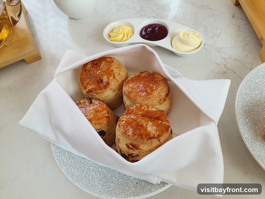 A basket of four golden-brown scones, two plain and two with visible raisins, is presented on a white cloth. Beside the basket, a small white dish holds dollops of butter and jam. A teapot and a patterned plate are also visible on the table.