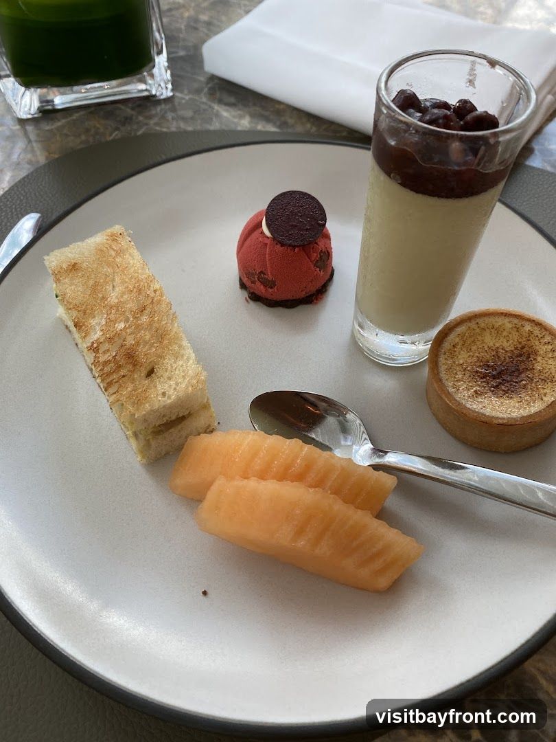 A plate with a selection of desserts and fruit. Items include a small red dome-shaped cake with a chocolate disc on top, a glass of panna cotta topped with red beans and a dark sauce, a small tart with a brûléed top, slices of cantaloupe, and a small sandwich. A spoon rests on the plate. A glass of green liquid is visible in the upper left corner.