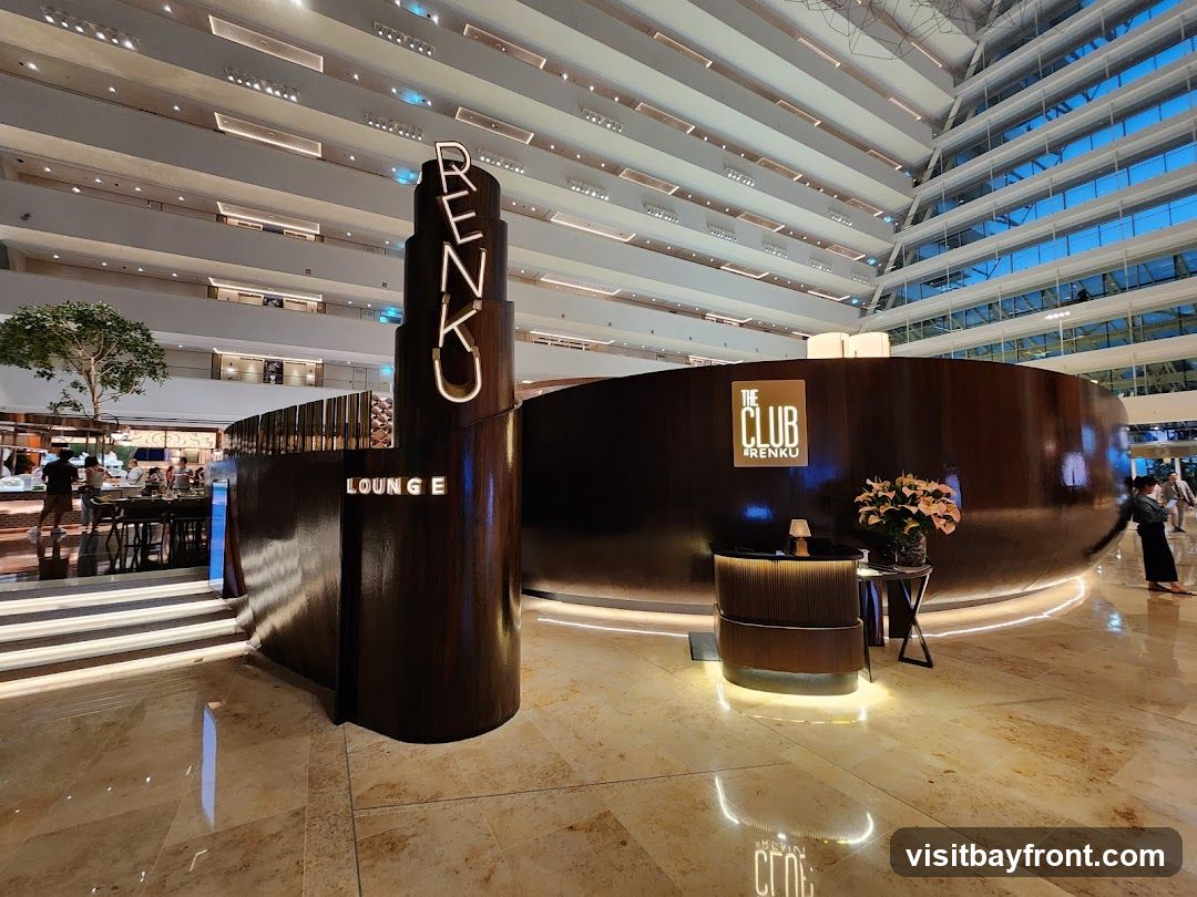 A modern, upscale lounge area with a curved wooden reception desk and a tall, illuminated sign that spells out "RENKU". The lounge is situated within a large, multi-level atrium with a distinctive sloped ceiling and abundant natural light. In the background, patrons can be seen at a bar area.