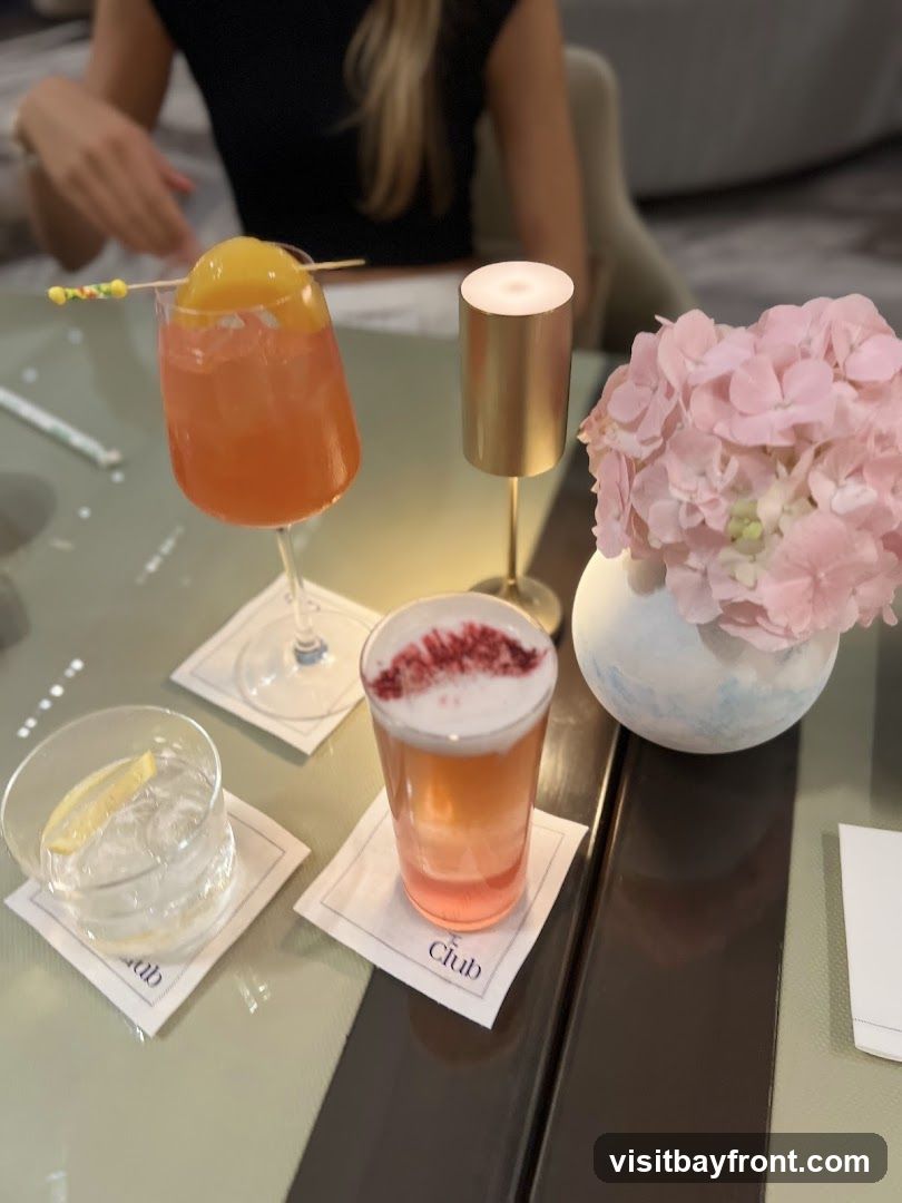 Three drinks are arranged on a table, with a vase of pink hydrangeas and a small lamp. One drink is in a stemmed glass and topped with a slice of peach. Another is in a shorter glass with a foamy top dusted with red berries. The third is a clear drink with ice and a lemon slice. The coasters under two of the drinks have the word "Club" printed on them. A person's hands are visible in the background.