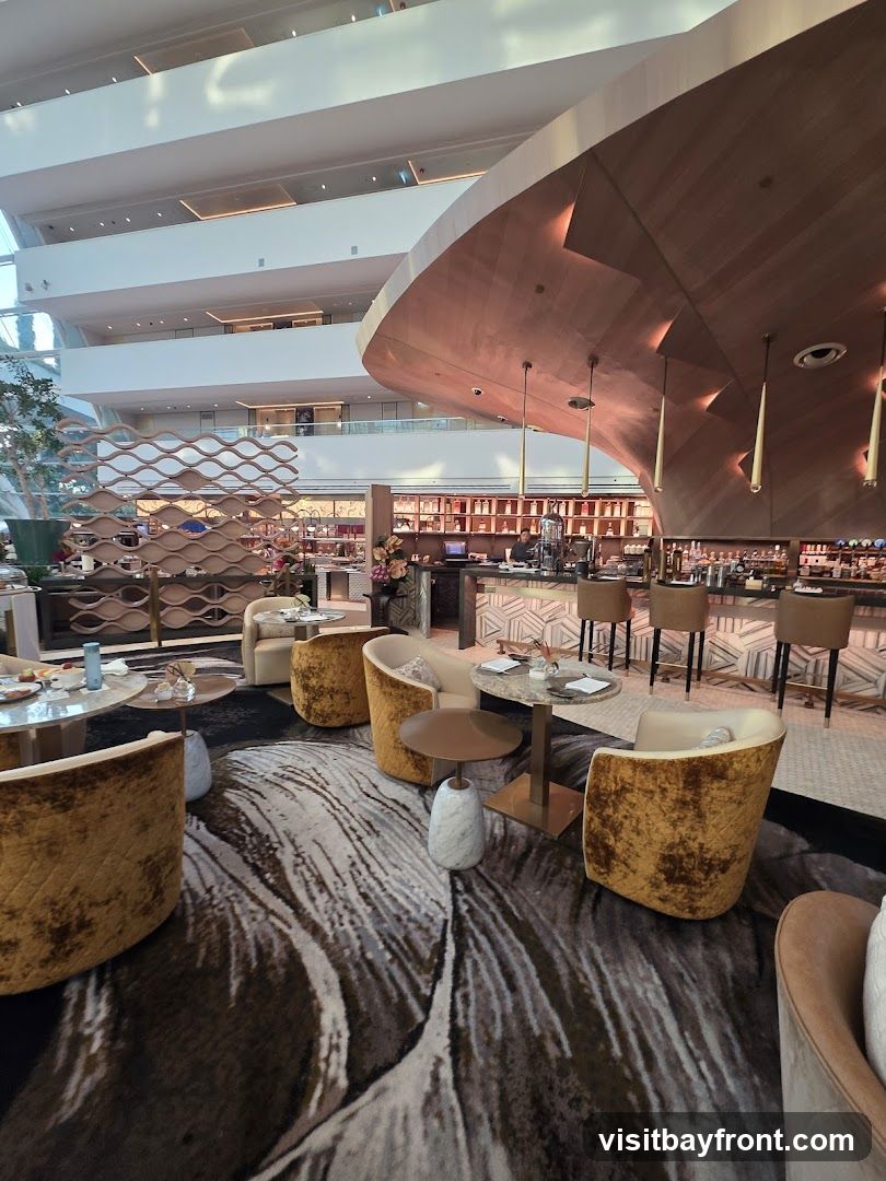 A contemporary bar and lounge area with plush seating, marble-topped tables, and a distinctive curved wooden bar. The space features a large, abstract-patterned carpet and is set within a multi-level atrium with natural light.