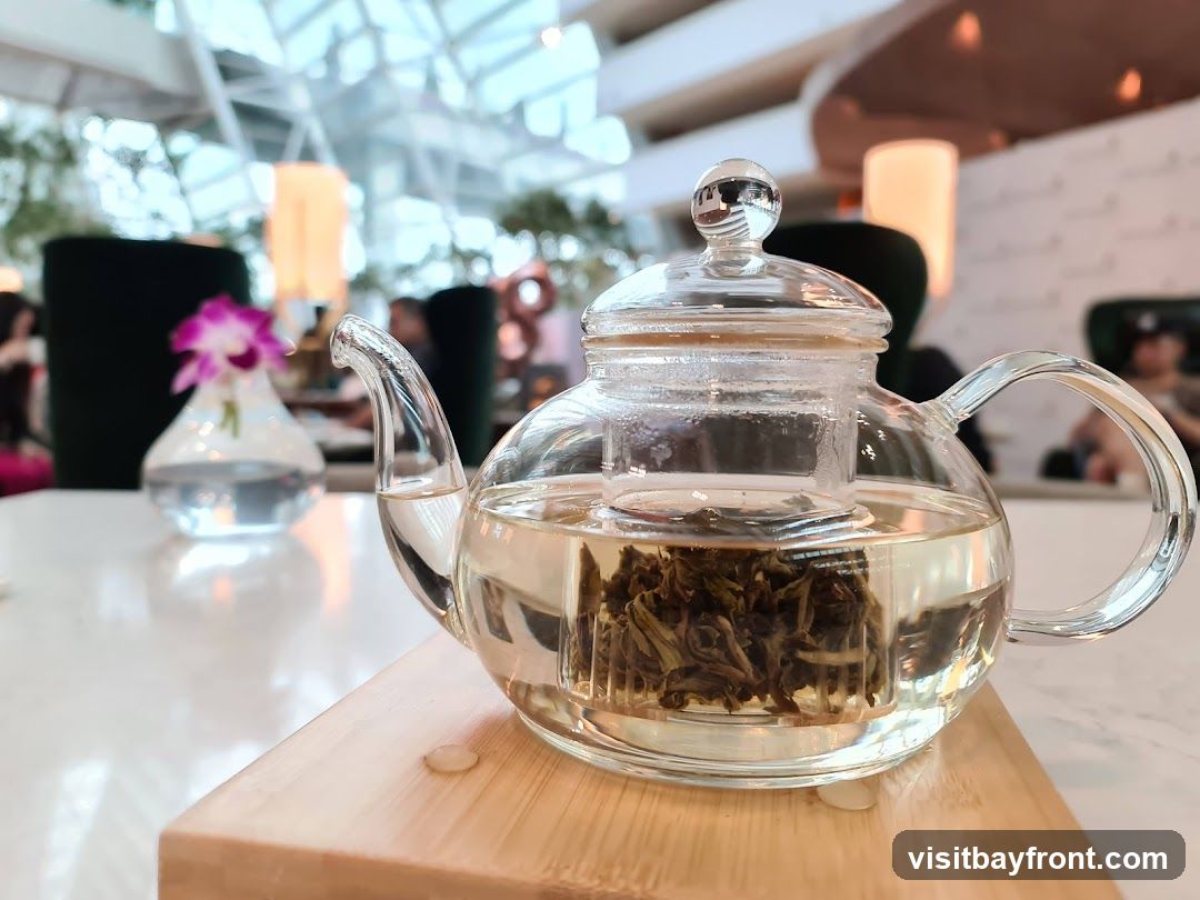 A clear glass teapot filled with hot water and loose leaf tea is placed on a wooden coaster on a white table. In the background, a small vase with a pink orchid and a blurred interior of a cafe or restaurant can be seen.