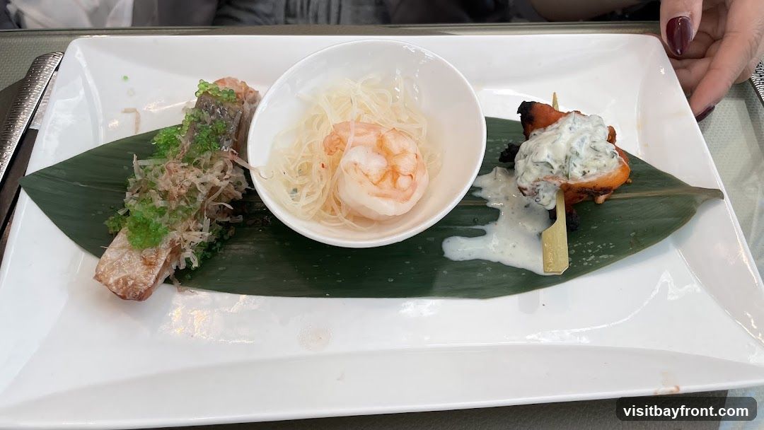 A white rectangular plate holds a trio of appetizers. On the left, a piece of grilled fish is topped with green roe and dried bonito flakes. In the center, a single shrimp sits in a white bowl filled with shredded noodles. On the right, a skewer of grilled meat is served with a dollop of creamy sauce. The appetizers are presented on a large green leaf.