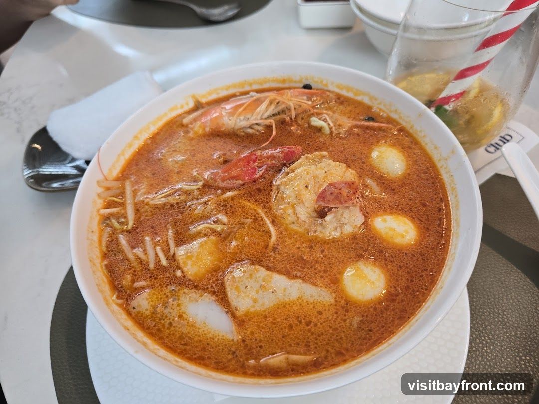 A close-up shot of a bowl of seafood noodle soup. The soup is a vibrant orange-red color and contains two large prawns, bean sprouts, and white fish cakes. A glass with a red and white striped straw and lemon slices is visible in the background, along with a white napkin and a spoon.