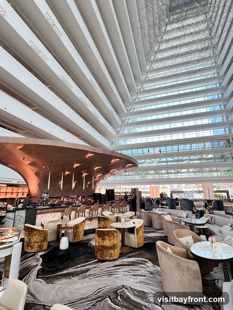 An interior view of a spacious, modern bar and lounge area with a high, multi-tiered ceiling and a striking architectural design. The space features a curved bar with pendant lighting, numerous seating arrangements including plush armchairs and sofas, and small tables. The decor is elegant, with a patterned carpet and warm-toned upholstery. Natural light streams in from the large, glass-enclosed atrium above.