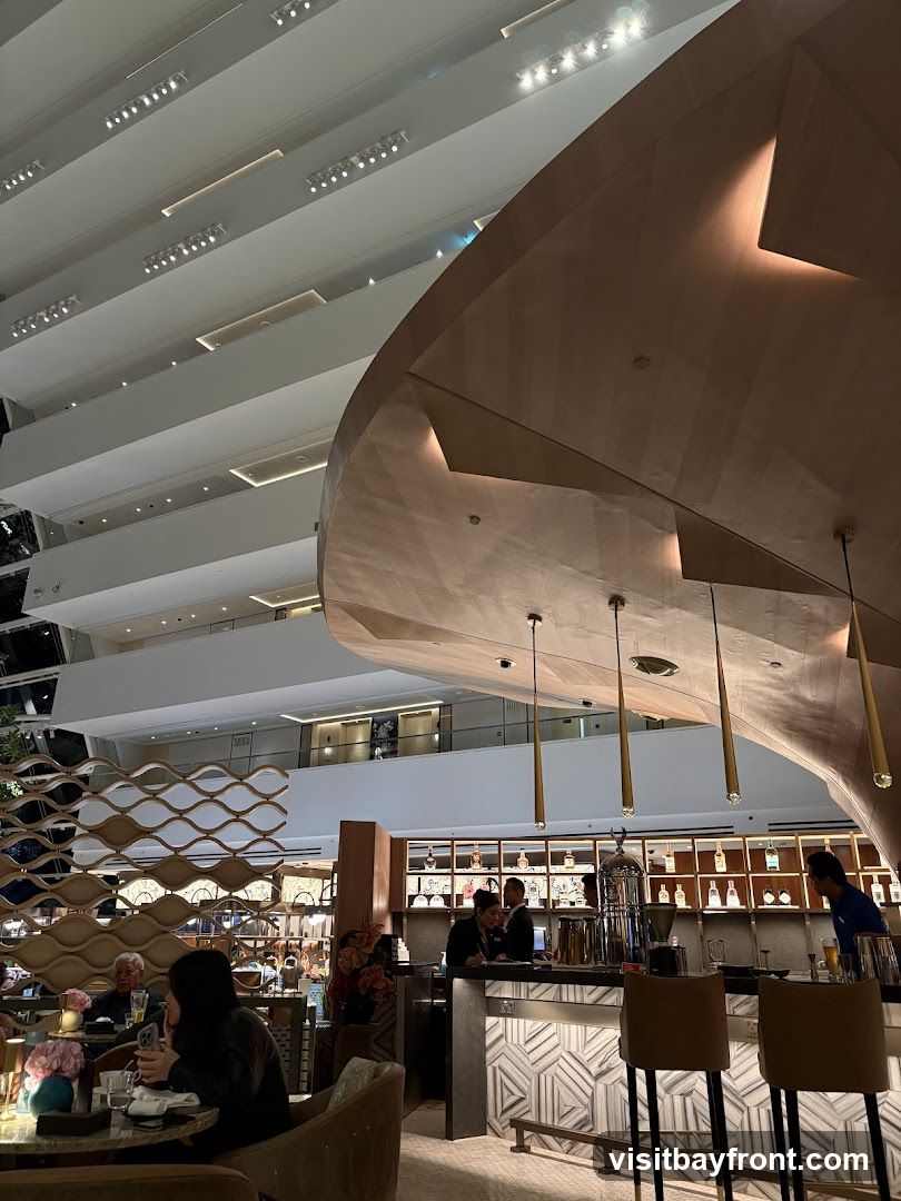 A dimly lit, upscale bar with a large, curved wooden structure overhead. Patrons are seated at tables and at the bar, where bartenders are serving drinks. The bar area features a well-stocked shelf of bottles and modern geometric tiling on the counter. The overall ambiance is sophisticated and contemporary.