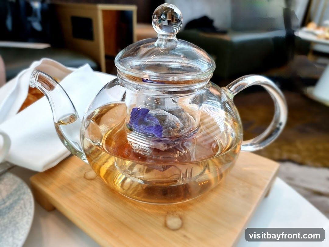 A clear glass teapot filled with amber-colored tea and a purple flower is placed on a wooden stand. The teapot has a glass lid with a knob and a glass handle. In the background, a white napkin and out-of-focus furniture are visible.