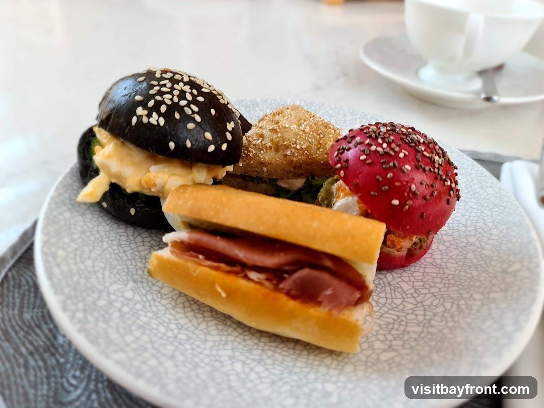 A close-up shot of a plate with three mini sandwiches and one long sandwich. One mini sandwich has a black bun topped with sesame seeds and appears to be filled with a creamy egg salad. Another mini sandwich has a bright red bun sprinkled with chia seeds and other seeds, with visible filling. A third mini sandwich has a light brown bun with seeds. The long sandwich is a baguette-style roll filled with thinly sliced meat, possibly ham. In the background, a white teacup and saucer are visible.