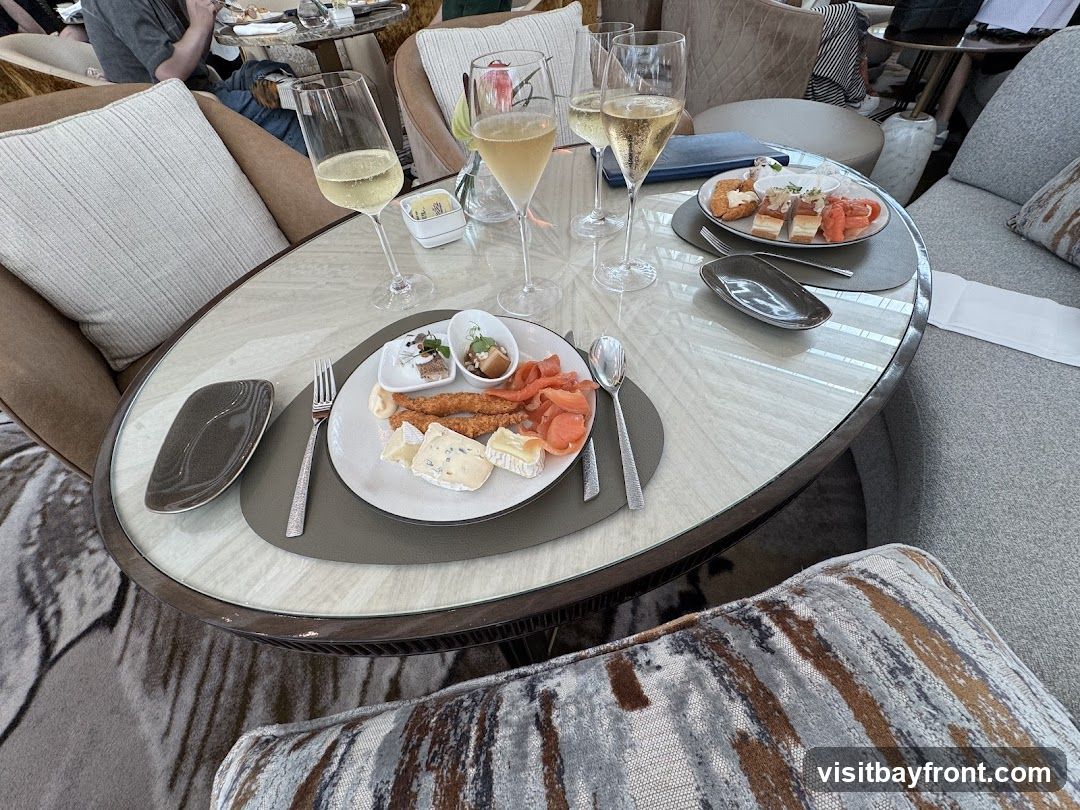 A table setting with a platter of cheese, smoked salmon, and small appetizers, accompanied by glasses of champagne. The table is oval with a marble-like surface and a dark rim. The seating appears to be plush armchairs and a sofa, suggesting a lounge or dining area.