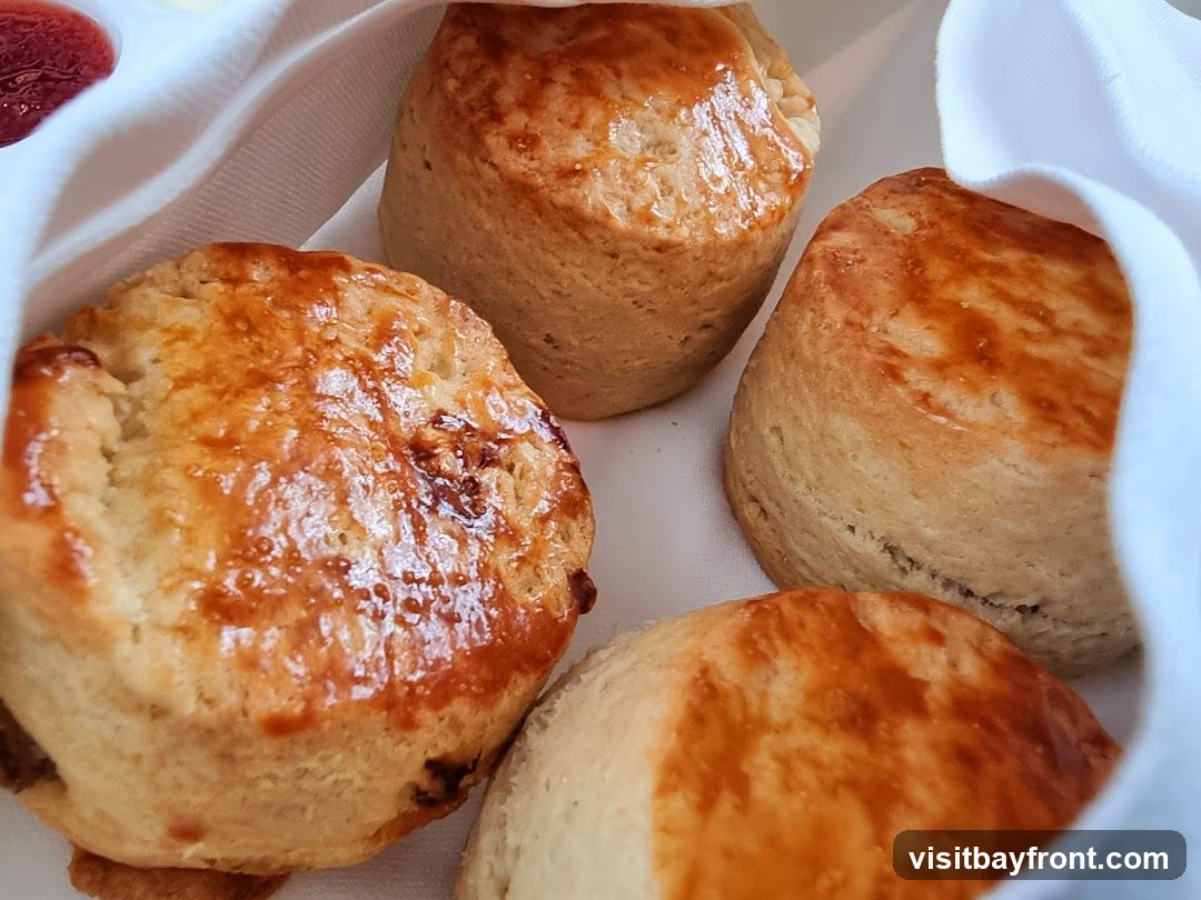 A close-up shot of four golden-brown scones, partially covered by a white cloth. A small dish of red jam and a dollop of butter are visible in the upper left corner.