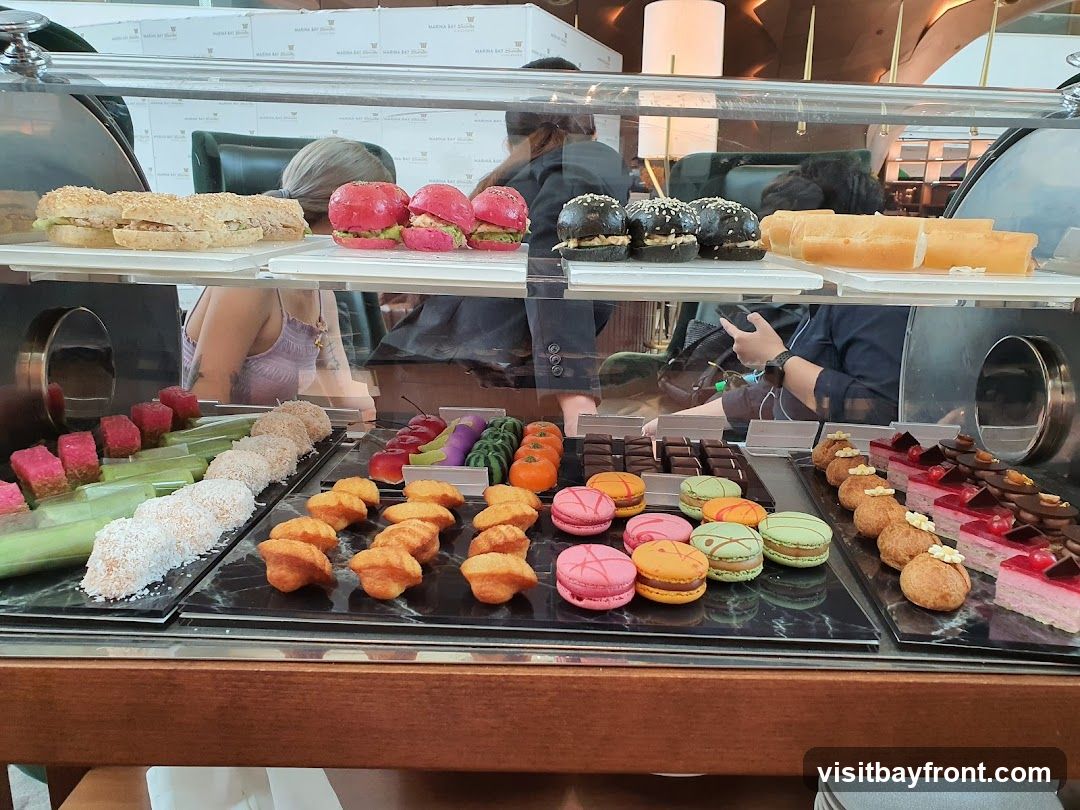 A display case filled with various desserts and sandwiches. On the top shelf, there are sandwiches, pink burgers, black burgers, and what appear to be bread rolls. The lower shelf features an assortment of colorful sweets including pink and green cubes, coconut-covered pastries, small cakes, macarons in various colors, eclairs, and layered cakes with fruit toppings.
