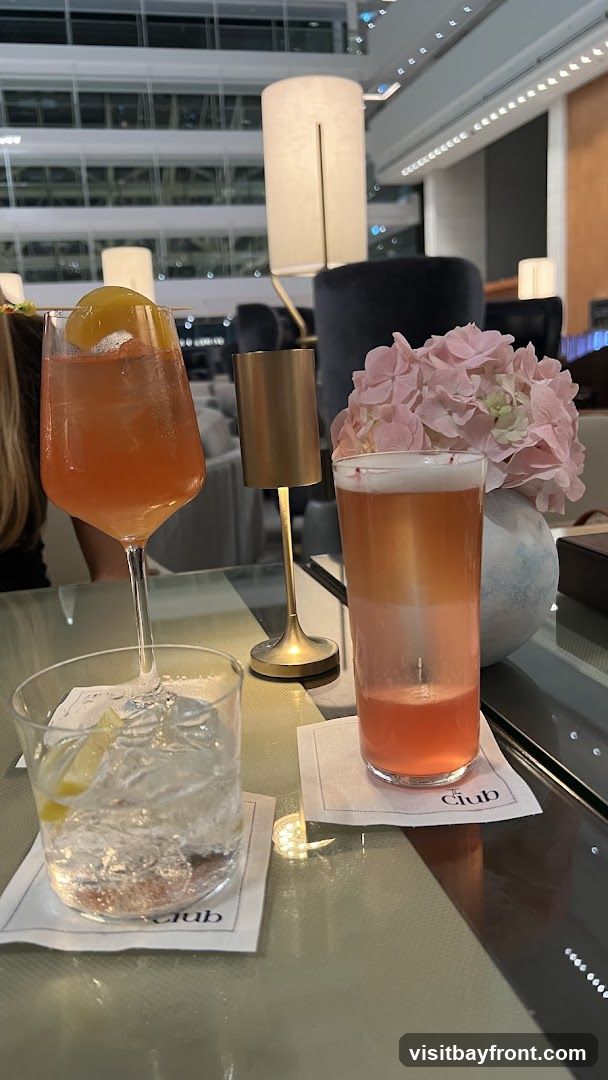 Three cocktails are arranged on a table. The cocktail on the left is in a stemmed glass and garnished with a slice of peach. The cocktail in the middle is in a tall glass and has a foamy top. The cocktail on the right is in a rocks glass with ice and a lemon wedge. The table is set with white napkins that have the word "Club" printed on them. In the background, there are lamps and a floral arrangement.