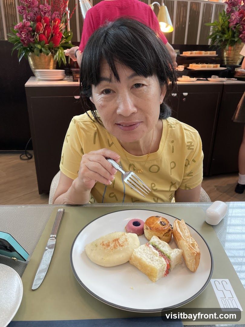 A woman in a yellow t-shirt sits at a table with a plate of food in front of her. The plate contains a sandwich, a pastry, a small pink dessert, and a savory item. She is holding a fork and appears to be about to eat. In the background, there is a buffet with more food items and floral arrangements. A logo with the text "55 CLUB" is visible on the table.
