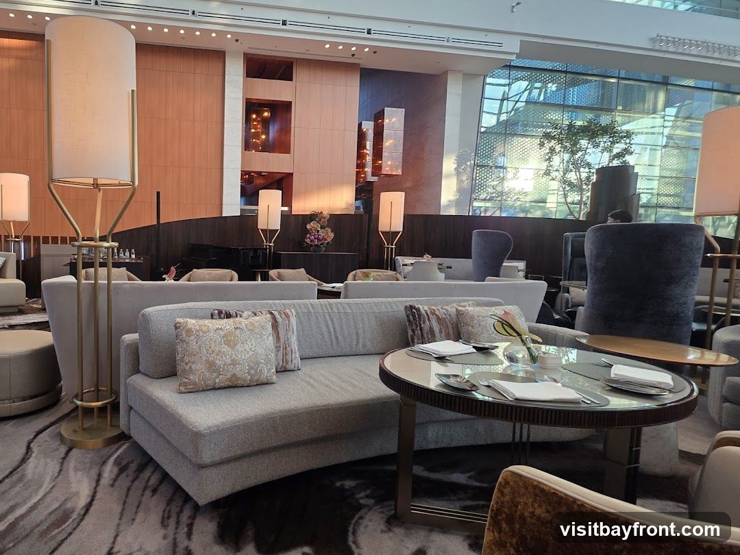 A luxurious and spacious lounge area with comfortable seating, elegant lighting, and modern decor. The seating arrangement includes plush sofas and armchairs, arranged around glass-topped tables set with napkins and cutlery. A grand piano is visible in the background, suggesting a sophisticated ambiance. Large windows offer a view of the outdoors, with sunlight streaming in and reflecting off the building's facade. The overall impression is one of refined comfort and style.