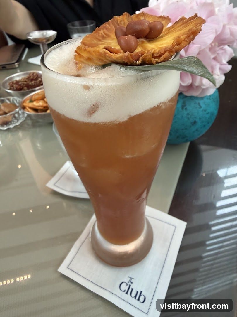 A tall glass of amber-colored cocktail topped with a thick layer of foam. The drink is garnished with a dried pineapple slice, several nuts, and a sage leaf. The glass sits on a white napkin with a blue logo that reads "The Club". In the background, there are small bowls of snacks, a vase with pink flowers, and a blue decorative object.