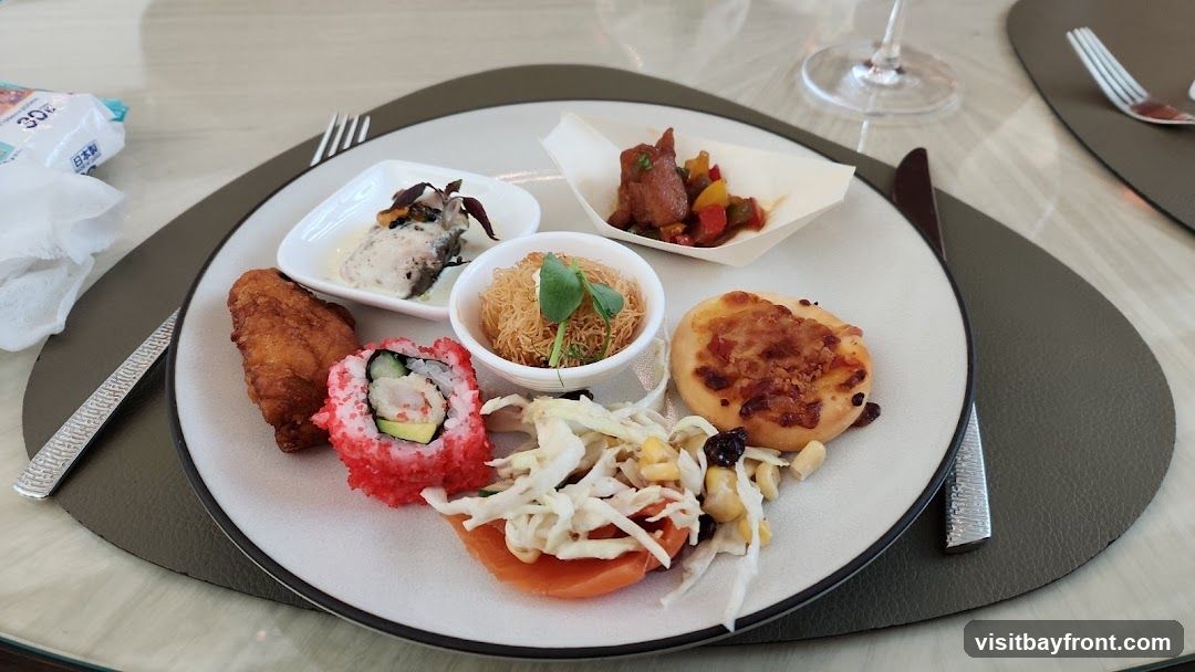 A plate of assorted appetizers is presented on a table. The plate contains a piece of fried chicken, a sushi roll with red roe, a small pizza, a salad with corn and raisins, a small bowl of crispy noodles, a dish with fish in sauce, and a small cup with meat and vegetables. A napkin and cutlery are also visible.