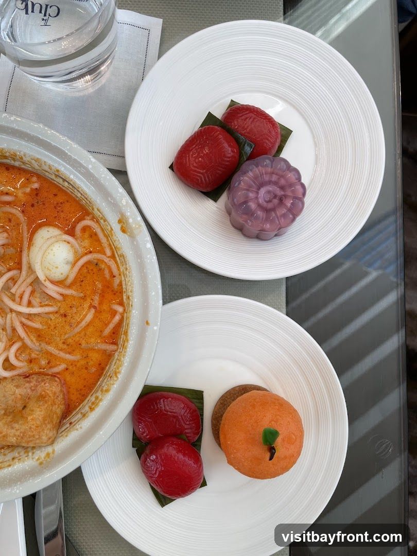 A bowl of laksa with noodles, egg, and a piece of fried tofu is shown next to two plates of colorful desserts. One plate has two red, intricately patterned pastries and a purple, flower-shaped jelly. The other plate has two similar red pastries, a round brown cookie, and an orange, pumpkin-shaped dessert with a green stem. A glass of water with a napkin bearing the logo "The Club" is also visible.