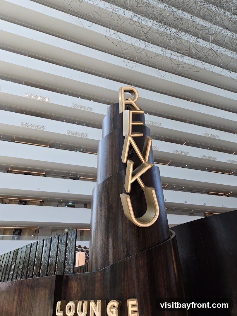 A tall, cylindrical wooden structure with illuminated golden letters spelling out "RENKU" is prominently displayed. Below this, the word "LOUNGE" is also visible in illuminated letters. The background shows multiple levels of a building interior with balconies and a modern, geometric ceiling installation.