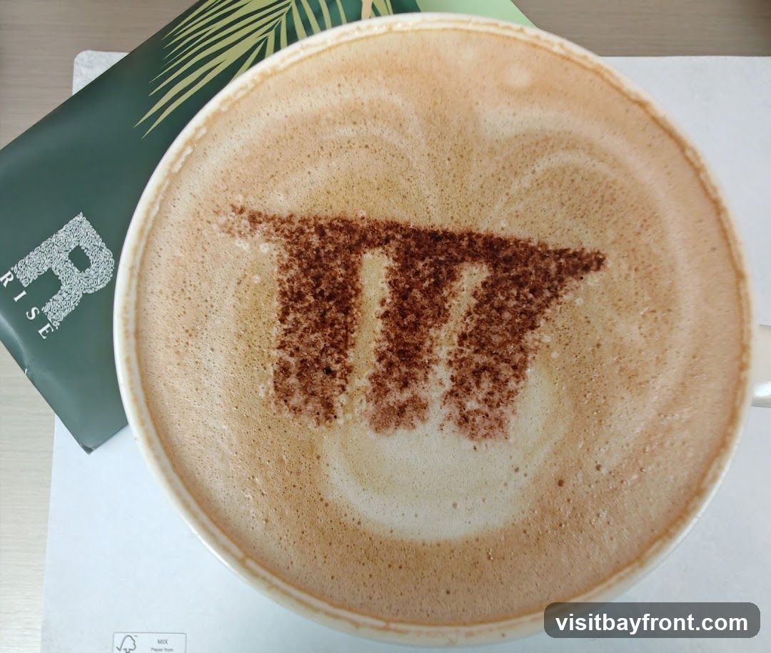 A close-up overhead view of a latte or cappuccino with a pi symbol dusted on top with cocoa powder. A green branded item with the letter R and a leaf design is visible to the left.
