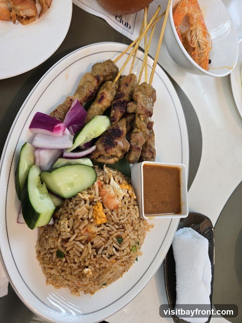 A plate of satay skewers served with fried rice, cucumber slices, red onion wedges, and a small dish of peanut sauce. A small bowl of shrimp is visible in the upper right corner. A folded white napkin is on a dark dish to the right of the plate.