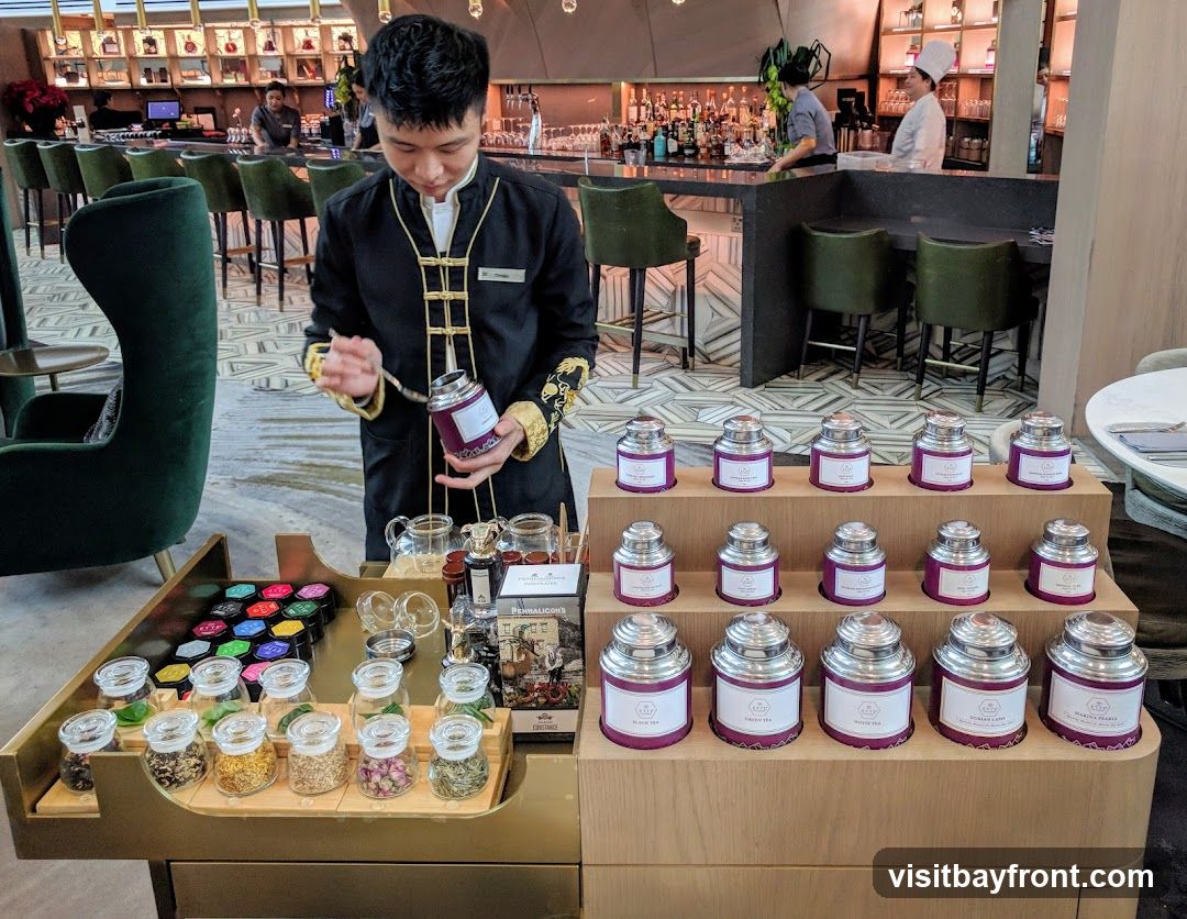 A server in a black uniform with gold trim presents a selection of teas. The display features rows of silver canisters with purple labels, and small glass jars containing various dried tea leaves and flower buds. In the background, a bar area with stools and bottles is visible, along with other staff members.
