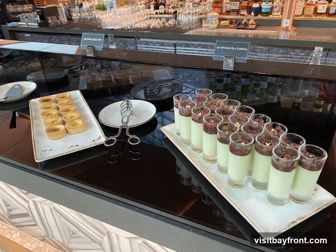 A display of desserts on a counter, with a bar in the background. In the foreground, there are small tarts and individual servings of matcha milk pudding. Labels indicate "HOKKAIDO CHEESE TART" and "MATCHA MILK PUDDING".