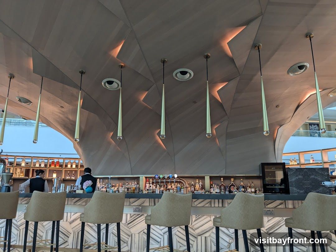 A contemporary bar with a striking, angular wooden ceiling and unique pendant lights. The bar itself is sleek, with a marble-patterned front and comfortable, upholstered stools. Behind the bar, bottles of liquor are neatly arranged on shelves, and two bartenders in uniform are visible.