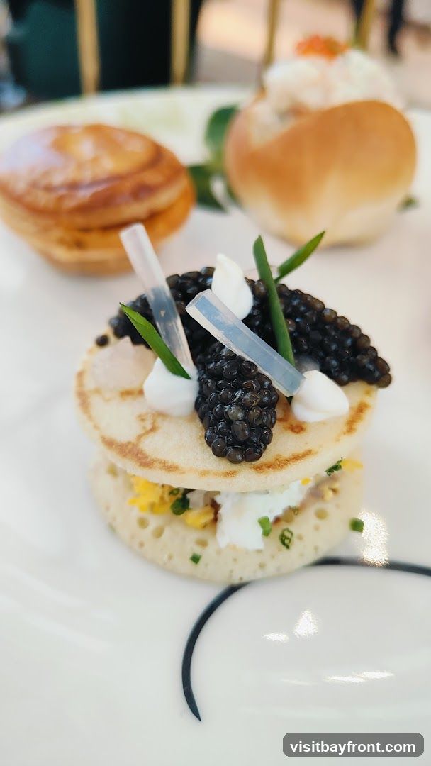 A close-up shot of blinis topped with caviar, cream, and chives. In the background, there are other small pastries and a bun with a filling. The food is served on a white plate with a dark curved design.