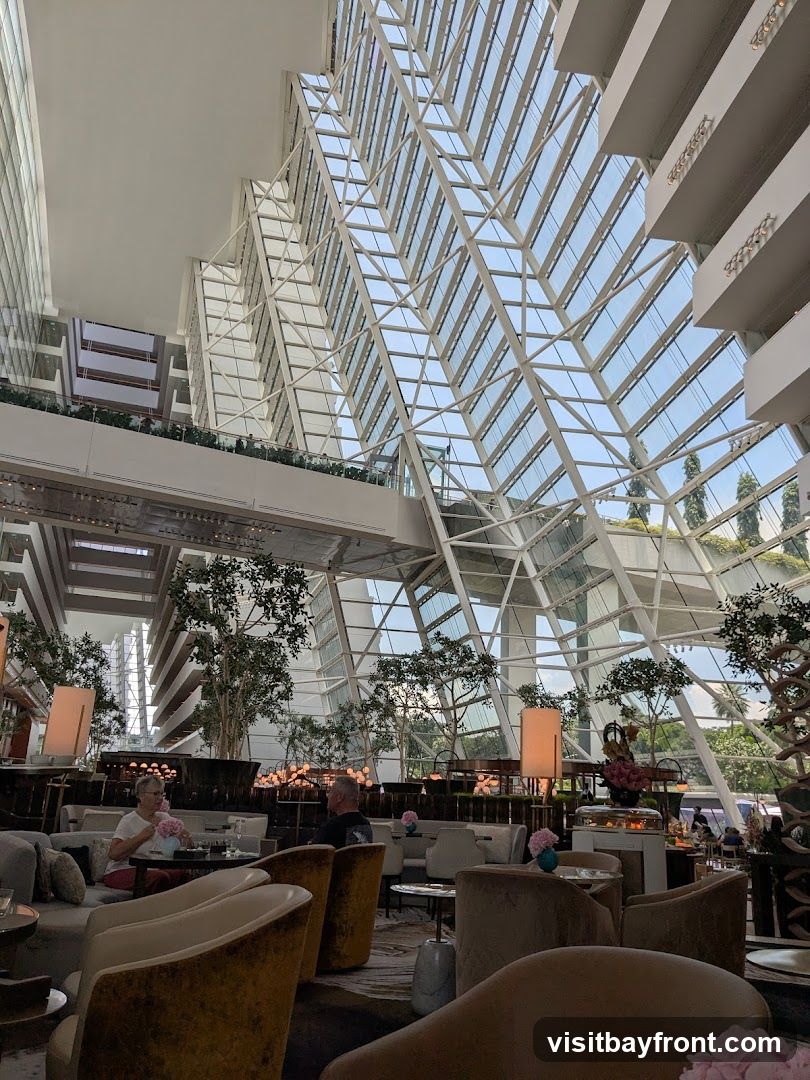 An interior view of a hotel atrium lounge with a large, geometric glass ceiling. Several levels of balconies with railings overlook the lounge area. Seating areas with plush chairs and sofas are arranged around tables. Potted trees and decorative lamps are placed throughout the space. Two people are seated at a table in the foreground.