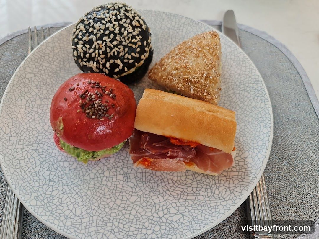 A plate holds four small food items: a dark bun covered in sesame seeds, a red bun topped with chia seeds and filled with green spread, a triangular pastry with a seeded crust, and a small baguette sandwich with ham and red sauce. The plate has a textured, crackled pattern. Cutlery is visible on either side of the plate.