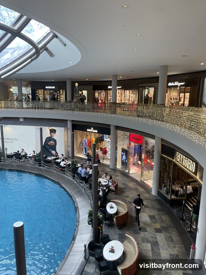 An interior view of a multi-level shopping mall with a large blue water feature in the foreground. Several shops are visible on the upper level, including BUCCELLATI, Salvatore Ferragamo, STONE ISLAND, and HUGO. There are also dining areas with people seated at tables. The mall is decorated with Christmas lights and trees.