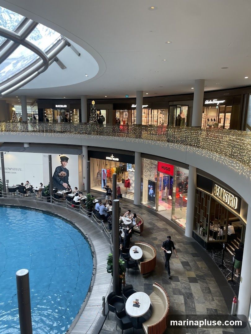 An interior view of a multi-level shopping mall with a large blue water feature in the foreground. Several shops are visible on the upper level, including BUCCELLATI, Salvatore Ferragamo, STONE ISLAND, and HUGO. There are also dining areas with people seated at tables. The mall is decorated with Christmas lights and trees.