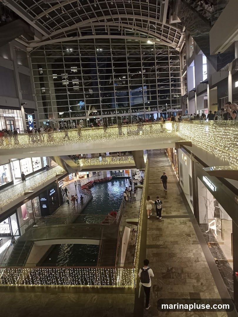 An interior view of a multi-level shopping mall featuring a canal with gondolas. The mall is decorated with numerous string lights, creating a festive atmosphere. People are seen walking on the elevated walkways and browsing in the shops. The architecture includes a large glass facade reflecting the interior and exterior lights.