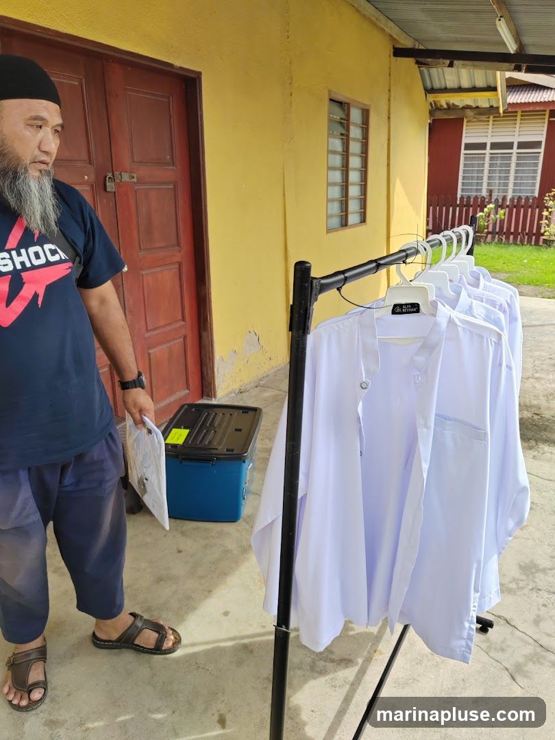 A man with a beard and a black cap stands next to a rack of white shirts. The shirts are hanging on hangers and appear to be traditional attire. The man is wearing a dark blue t-shirt and dark pants. Behind him is a yellow building with red doors and windows. A blue cooler box is also visible.