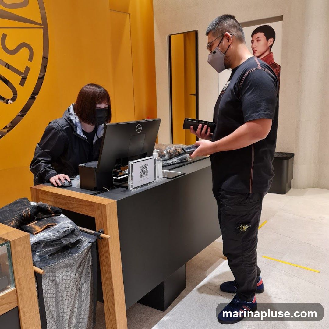 A person wearing a black jacket and mask is operating a computer at a checkout counter. Another person wearing a black t-shirt, mask, and black pants with a logo is standing at the counter holding a phone and a wallet. A QR code is displayed on a stand on the counter. A poster of a man is visible in the background.