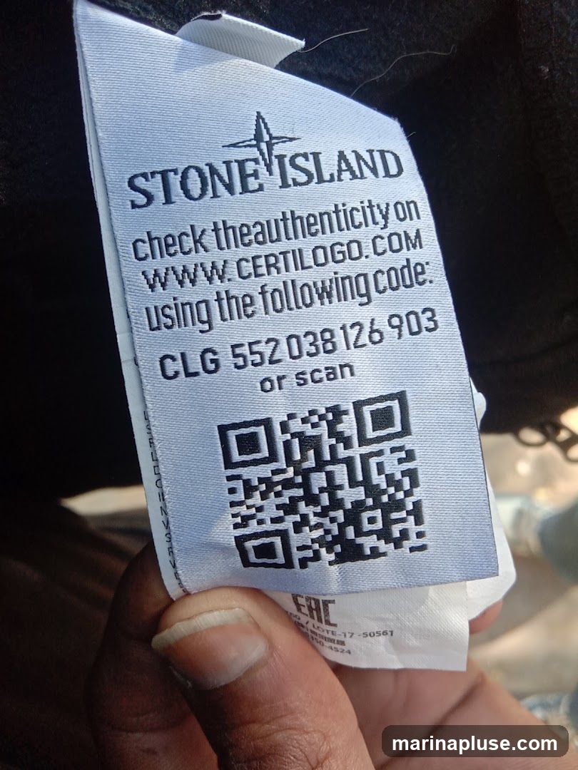 A close-up of a Stone Island clothing tag with instructions to check authenticity online via CERTILOGO.COM using a provided code or by scanning a QR code. The tag displays the Stone Island logo, the website, a code (CLG 552038126 903), and a QR code.