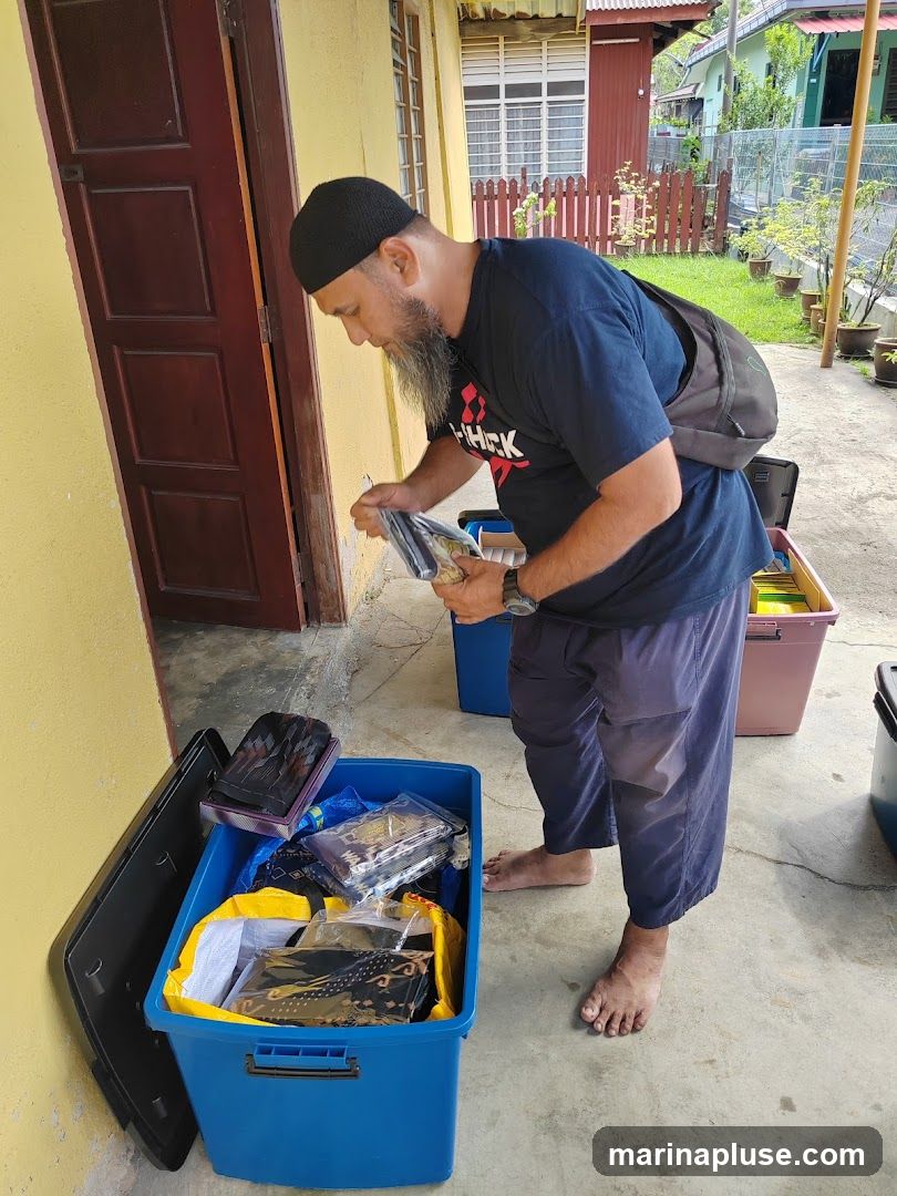 A man wearing a black skullcap and a dark t-shirt is standing barefoot outdoors, looking at items he is holding. He is surrounded by several large plastic bins filled with packaged goods, some of which appear to be textiles with intricate patterns. The bins are placed on a concrete surface next to a yellow building with a wooden door. In the background, there is a garden with a wooden fence and more houses.