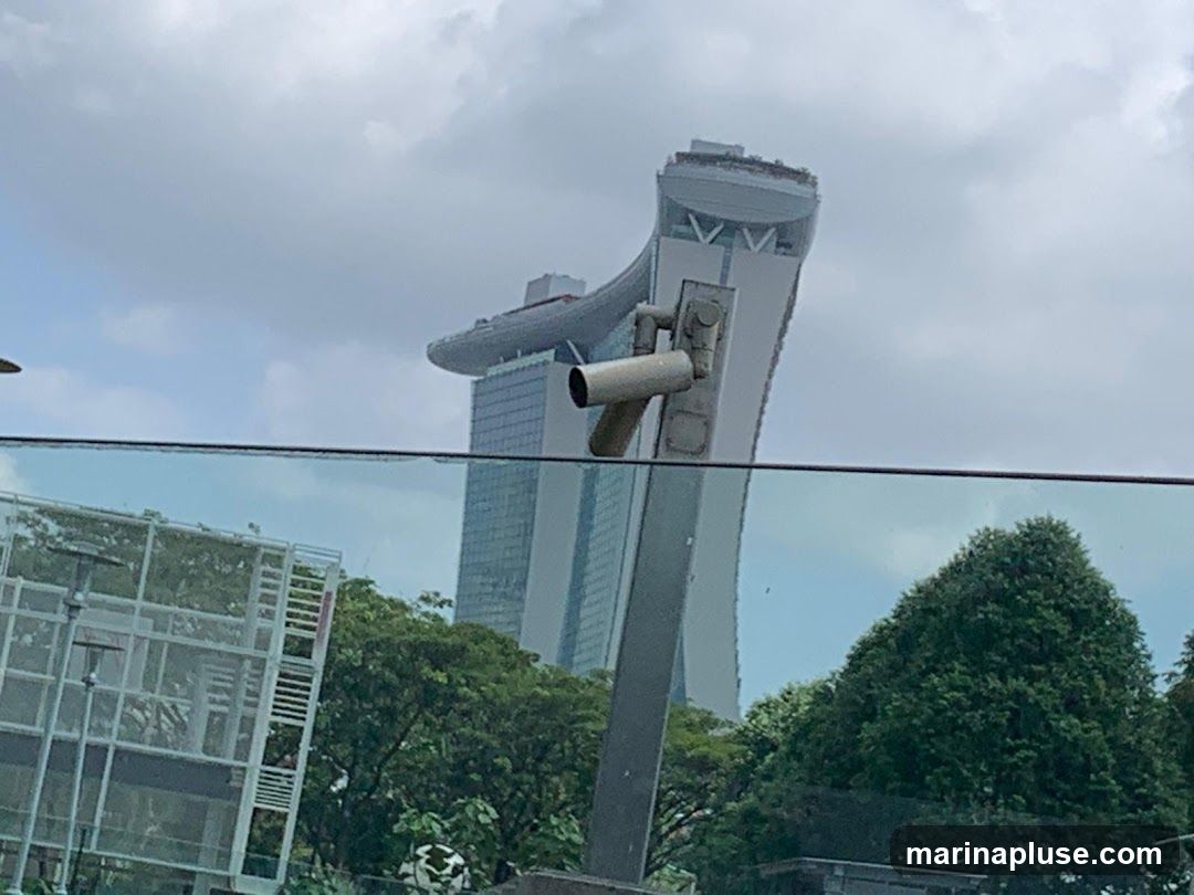 A view of the Marina Bay Sands building in Singapore under a cloudy sky. In the foreground, a glass barrier and some greenery are visible. A security camera is mounted on a pole in front of the building.
