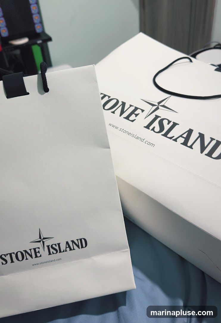 Two white Stone Island shopping bags are shown, one in the foreground and another slightly behind it. Both bags feature the Stone Island logo and website address printed in black. The bags appear to be made of paper and have black rope handles. The background is out of focus, showing a blue surface and a glimpse of a black object with colorful buttons.