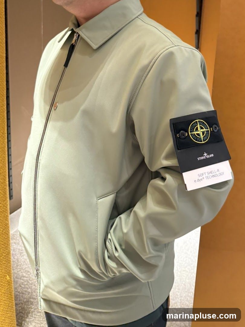 A person wearing a light green Stone Island jacket. The jacket has a zipper closure and a collar. A black patch with the Stone Island logo is visible on the left sleeve. A white tag with text is attached to the patch.