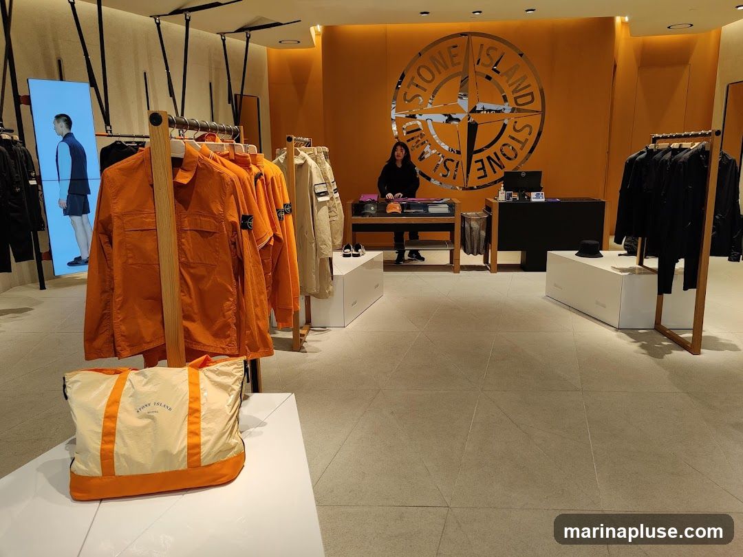 The interior of a Stone Island store features racks of clothing, including orange jackets and black garments. A large Stone Island logo is displayed on the orange wall behind a counter where a staff member is visible. A digital display shows a model wearing a vest and shorts. A tote bag with the Stone Island logo is placed on a white display stand in the foreground.
