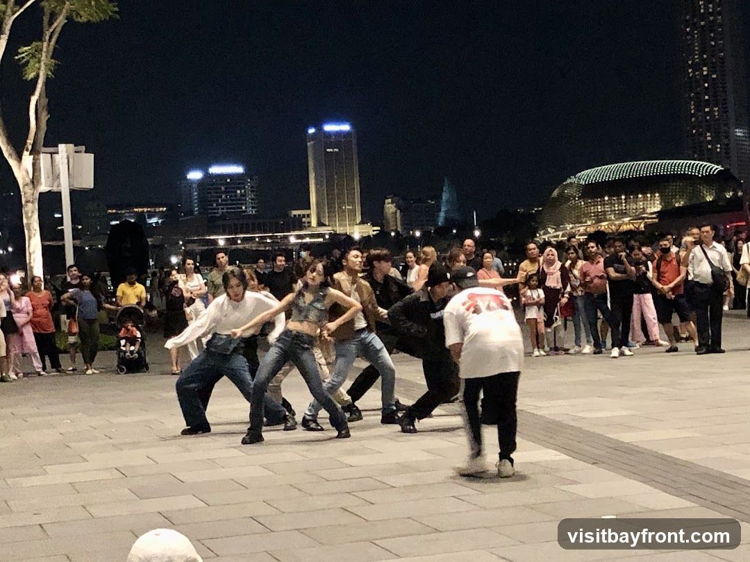 A group of dancers perform on a paved plaza at night, with a crowd of spectators watching. In the background, illuminated buildings and a distinctive domed structure are visible.