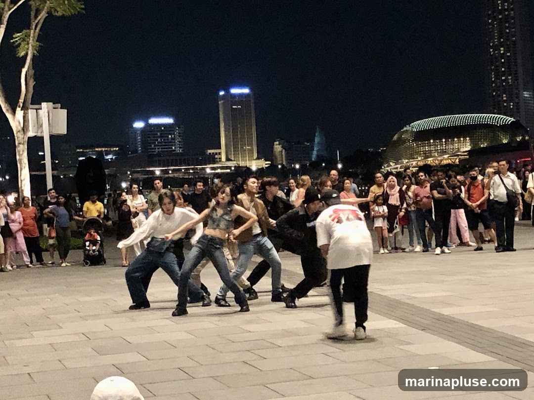 A group of dancers perform on a paved plaza at night, with a crowd of spectators watching. In the background, illuminated buildings and a distinctive domed structure are visible.