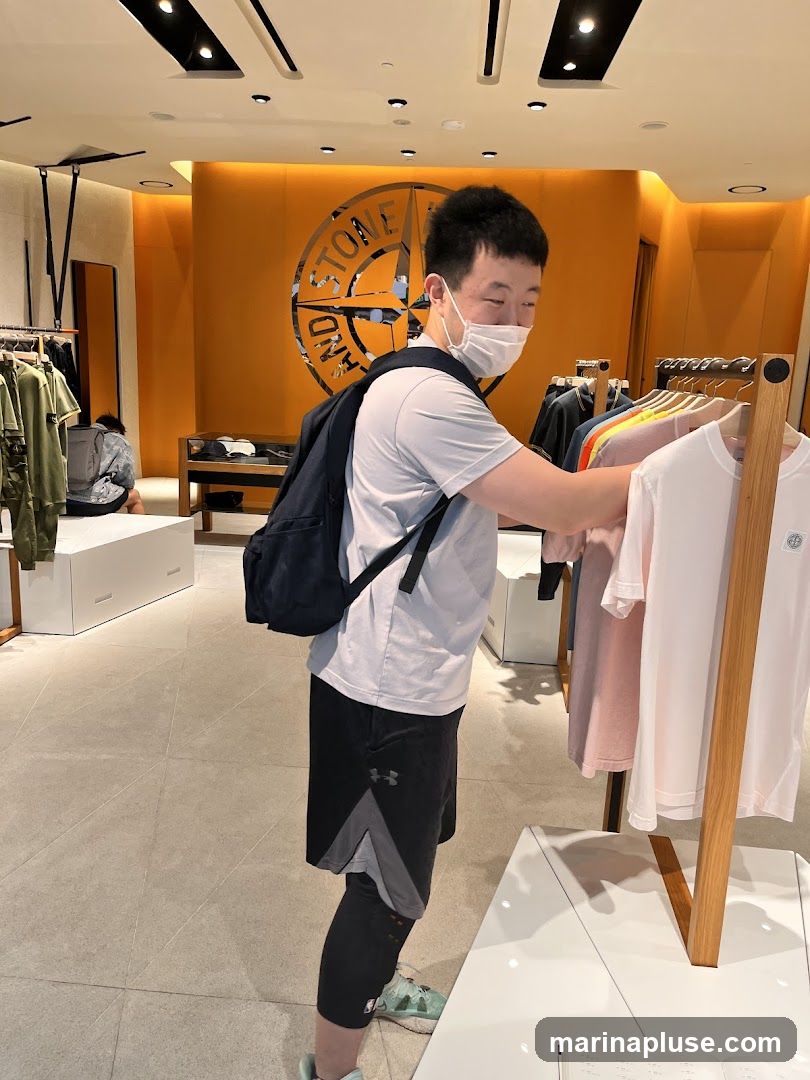 A man wearing a face mask and a backpack is browsing t-shirts on a rack in a brightly lit store. Behind him, a large circular logo with the words "STONE ISLAND" is visible on an orange wall. Racks of clothing are displayed throughout the store.