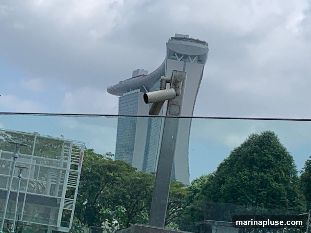 A tall, modern skyscraper with a distinctive sky bridge connecting its upper floors is visible against a cloudy sky. In the foreground, a security camera is mounted on a pole, and a glass barrier reflects trees and a building.