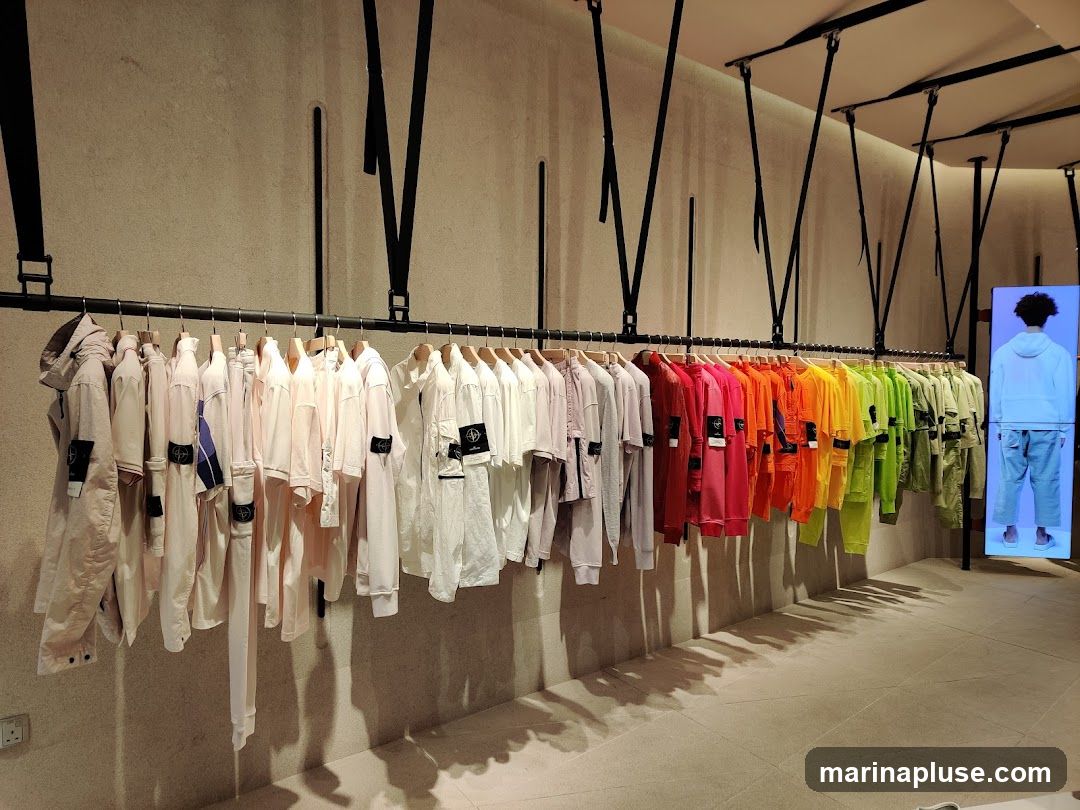 A clothing store display featuring a rack of garments in a gradient of colors from light pink to green. The garments appear to be shirts and jackets, many with visible brand logos. To the right, a digital screen displays a person wearing a white hoodie and pants.